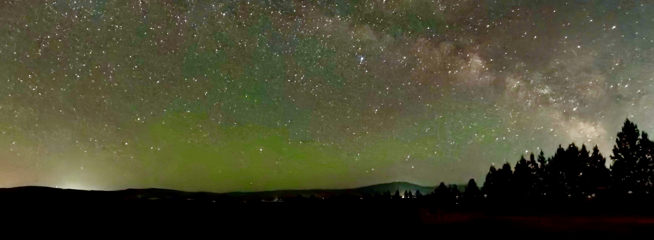 Panoramic Milky Way arching over evergreens with emerald green airglow and golden moonrise